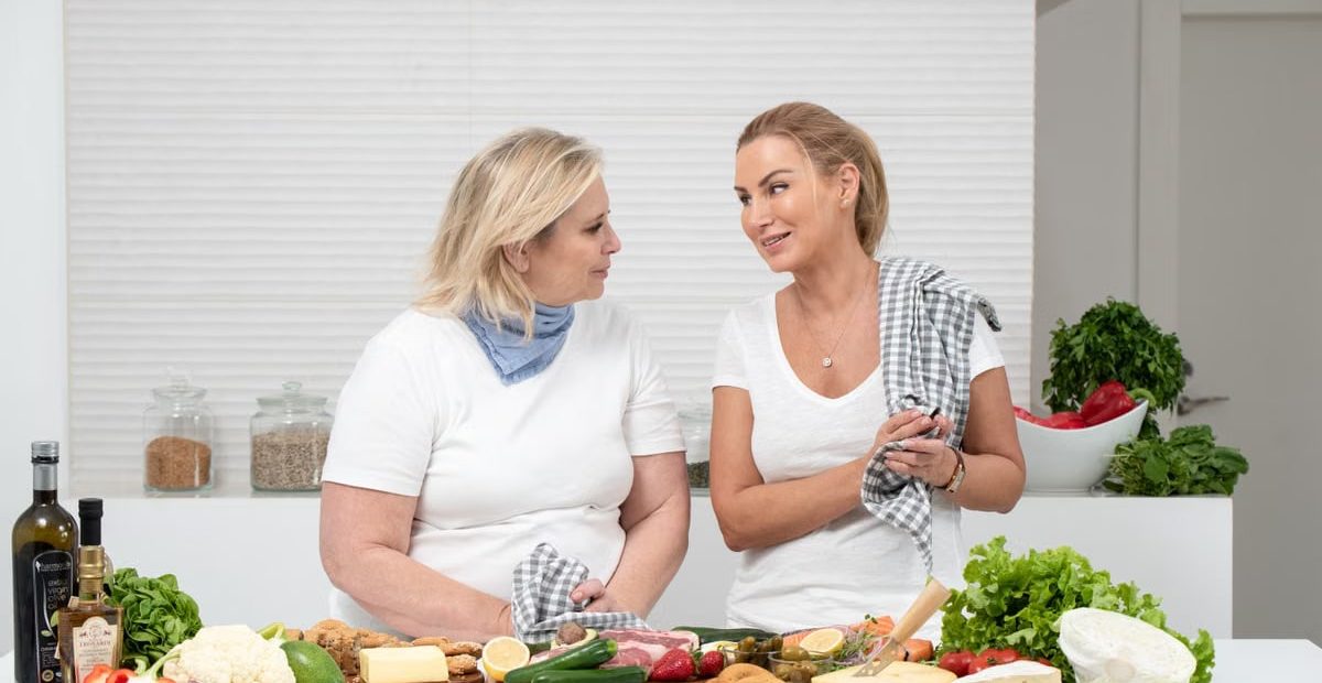 Two women in a kitchen preparing fresh vegetables and cheese with olive oil and spices.